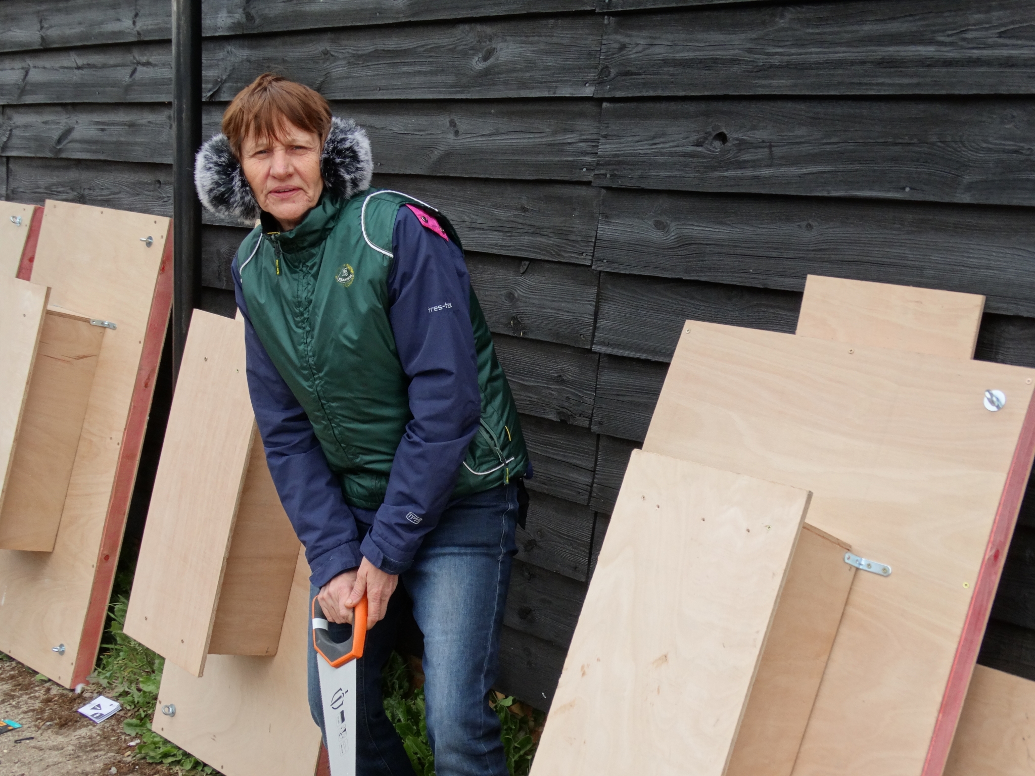 A photo from the FoTF Conservation Event - April 2019 - Mink Raft Construction at Santon Downham Workshops : A volunteers models a saw whilst partially constructed mink rafts stand up against the workshop in the background