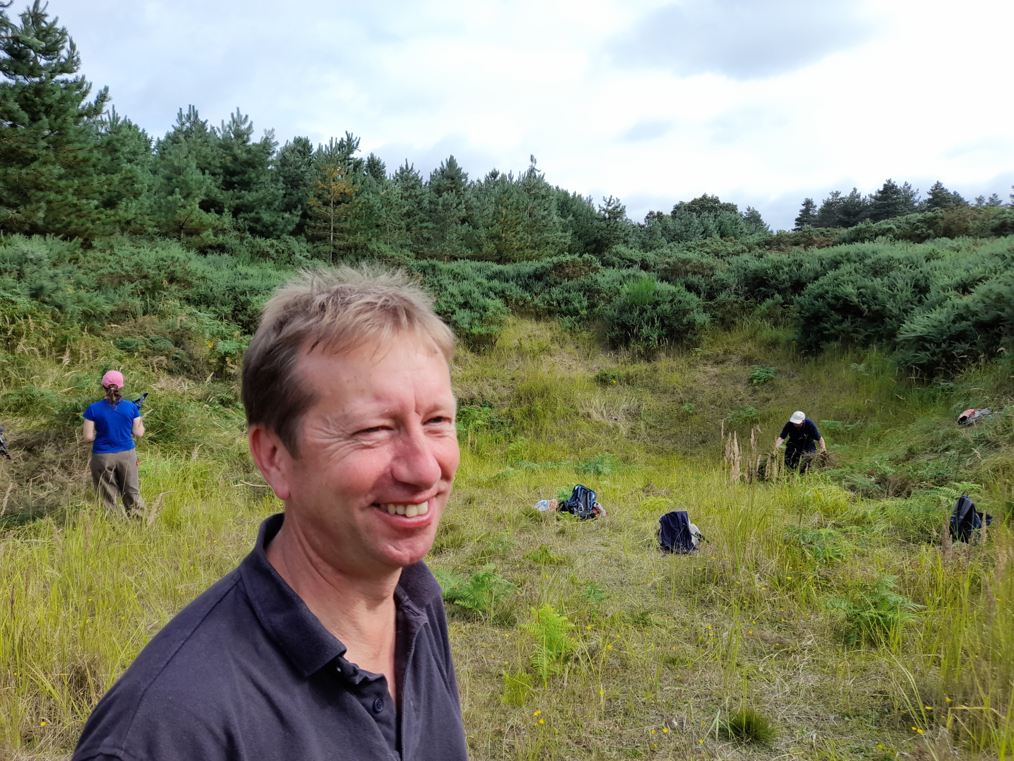 A photo from the FoTF Conservation Event - August 2021 - Maintenance tasks at Mildenhall Mugwort Pit & Mildenhall Warren Lodge : A volunteers smiles for the camera, while other volunteers work in Mildenhall Mugwort Pit in the background